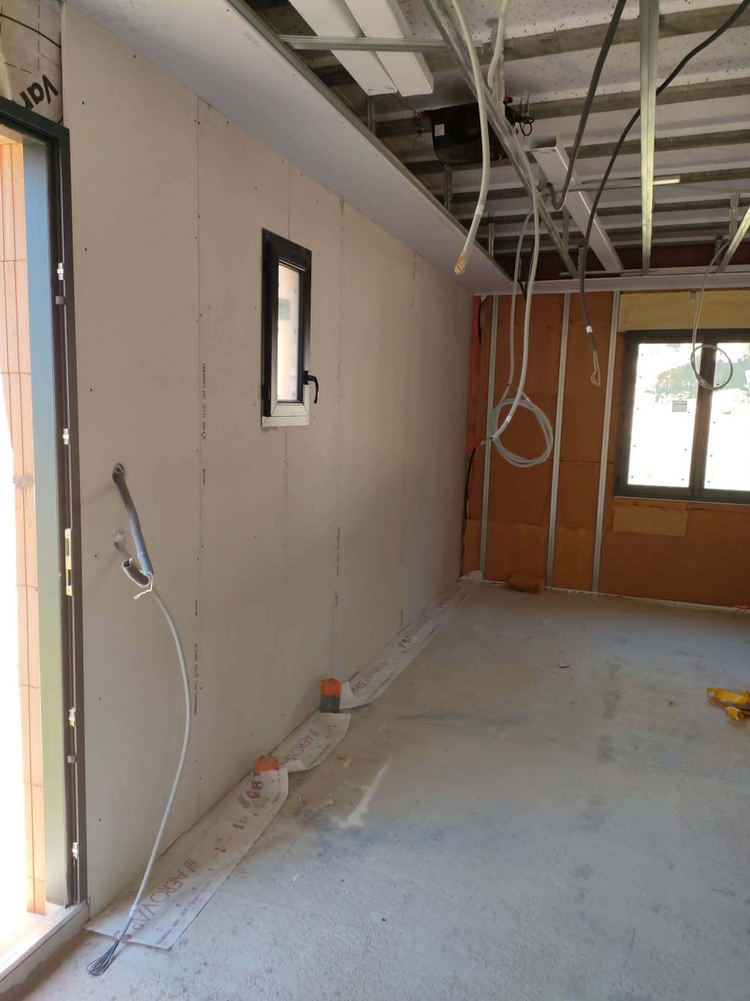 Mur recouvert de plaques de plâtre avec ossature de plafond et gaines électriques visibles dans une pièce en chantier.