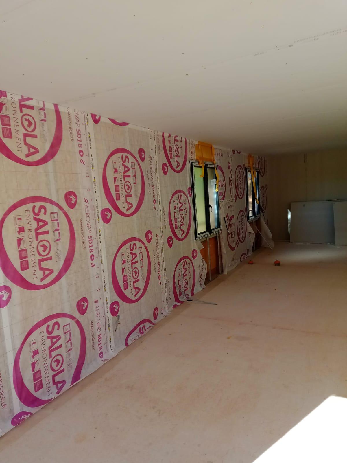 Murs recouverts d'un isolant visible dans une pièce en travaux, avec deux fenêtres et un plafond terminé.