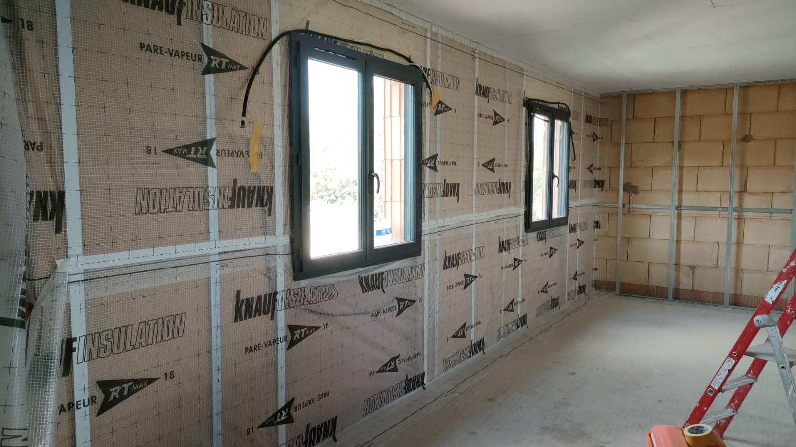 Pièce en chantier avec isolation et pare-vapeur visibles sur les murs, fenêtres posées et ossature métallique apparente.