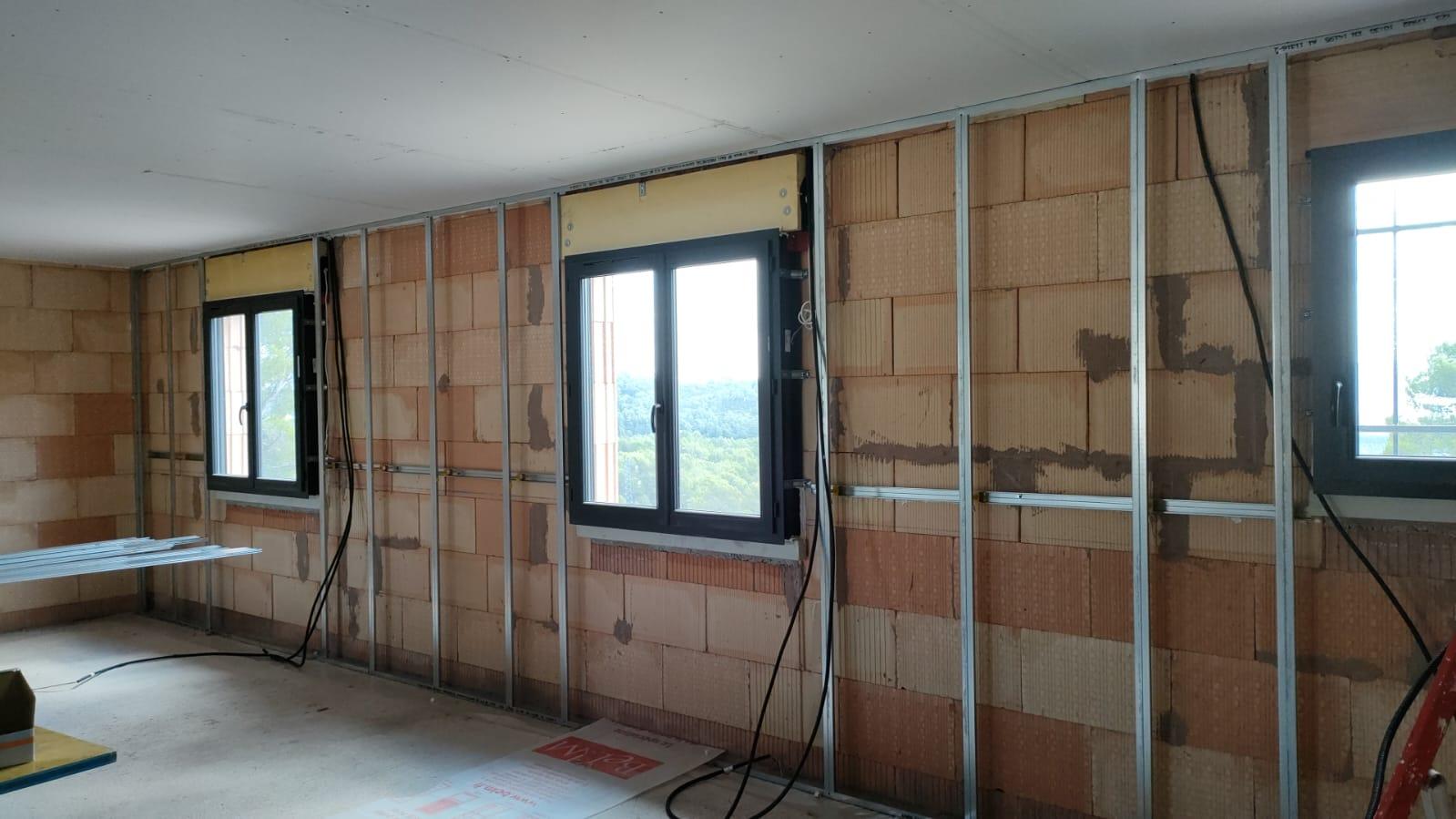 Ossature métallique installée devant un mur en briques avec plusieurs fenêtres dans une pièce en chantier.