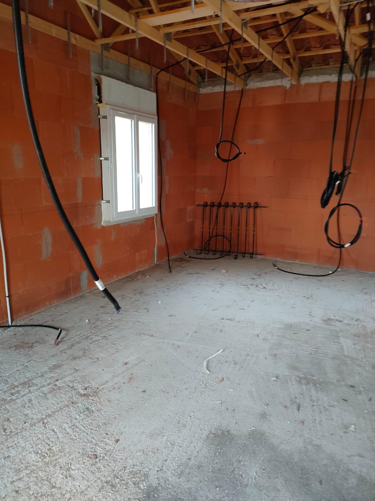 Pièce intérieure en chantier avec murs en briques apparentes, fenêtre, câbles et conduites visibles sous une charpente en bois.