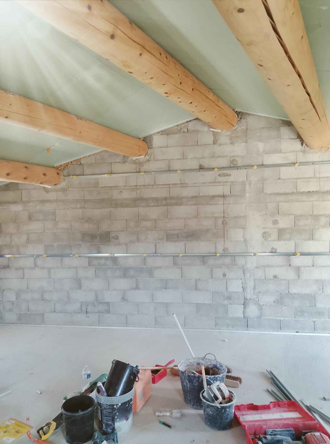 Plaques de plâtre posées au plafond entre des poutres en bois, avec des rails métalliques fixés sur un mur en blocs.